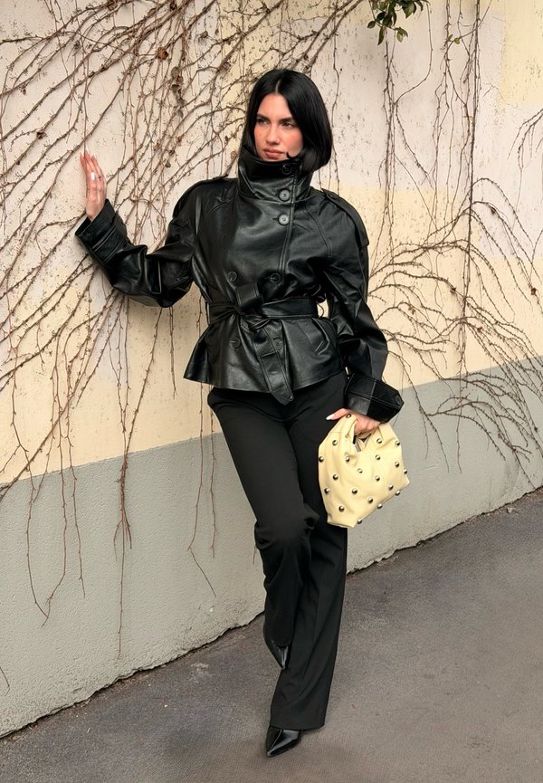 Γυναίκα με μαύρο δερμάτινο μπουφάν και παντελόνι στηρίζεται σε τοίχο καλυμμένο με κισσό, κρατώντας μια κρεμ τσάντα χειρός με καρφιά, στέκεται σε πλακόστρωτο έδαφος.