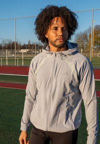 Helehall sportlik jope kapuutsiga, millel on peegeldavad detailid ja hingavust tagavad võrgust paneelid. Kantud mustade retuuside peal.