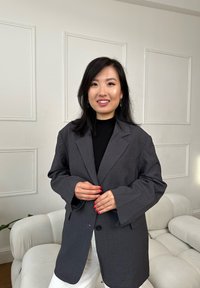 Blazer gris surdimensionné avec un seul bouton, manches larges et une poche. Porté sur un pull noir, associé à un pantalon blanc.