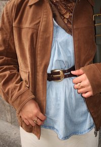 Braune Wildlederjacke mit Reißverschluss, hellblaues Denim-Oberteil, braigner Gürtel mit goldener Schnalle, gemusterter Schal und goldene Ringe an der Hand.