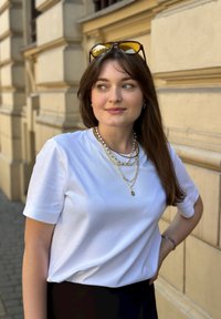 Ein weißes kurzärmeliges T-Shirt mit lässiger Passform, geschichtete Gold- und Perlenketten, runde Sonnenbrillen auf dem Kopf, posiert vor einer strukturierten Wand.