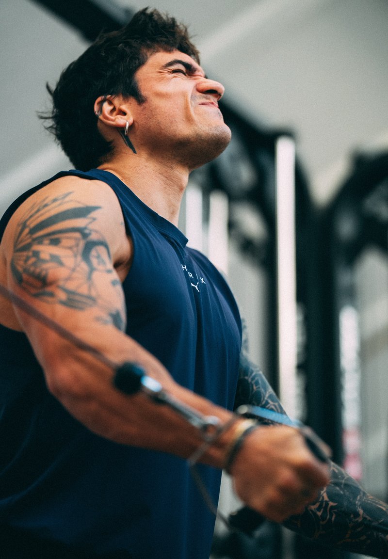 Homme avec des tatouages et des boucles d'oreilles qui se concentre en tirant un câble lors d'un entraînement de force dans une salle de sport.