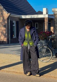 Μπλε παλτό καμπούρας πάνω από ένα πράσινο πλεκτό καζί, συνδυασμένο με μαύρα παντελόνια φαρδιάς γραμμής και μπεζ λόφους, αξεσουαρ ντυμένο με ακουστικά.