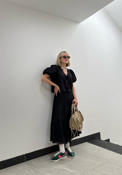 Top negro con mangas abullonadas y falda negra de volantes. Zapatillas verdes y rosas de Nike, calcetines blancos y un bolso beige con asas. Fondo minimalista.