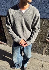 Pull gris à col en V en tissu maille doux, associé à un jean large bleu clair, et des chaussures noires, mains jointes à la taille.