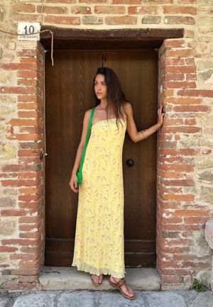Vestido maxi amarillo con un escote recto, acentuado por un bolso cruzado verde. Presenta una tela ligera y fluida, y sandalias planas.