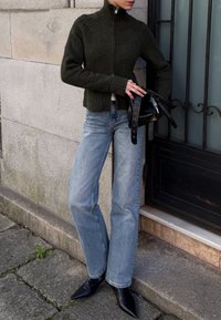 Pessoa a vestir um casaco de fecho éclair verde-escuro, jeans azul-claro e botas pretas de bico, segurando uma mala de couro preta junto a uma parede de pedra e uma porta.