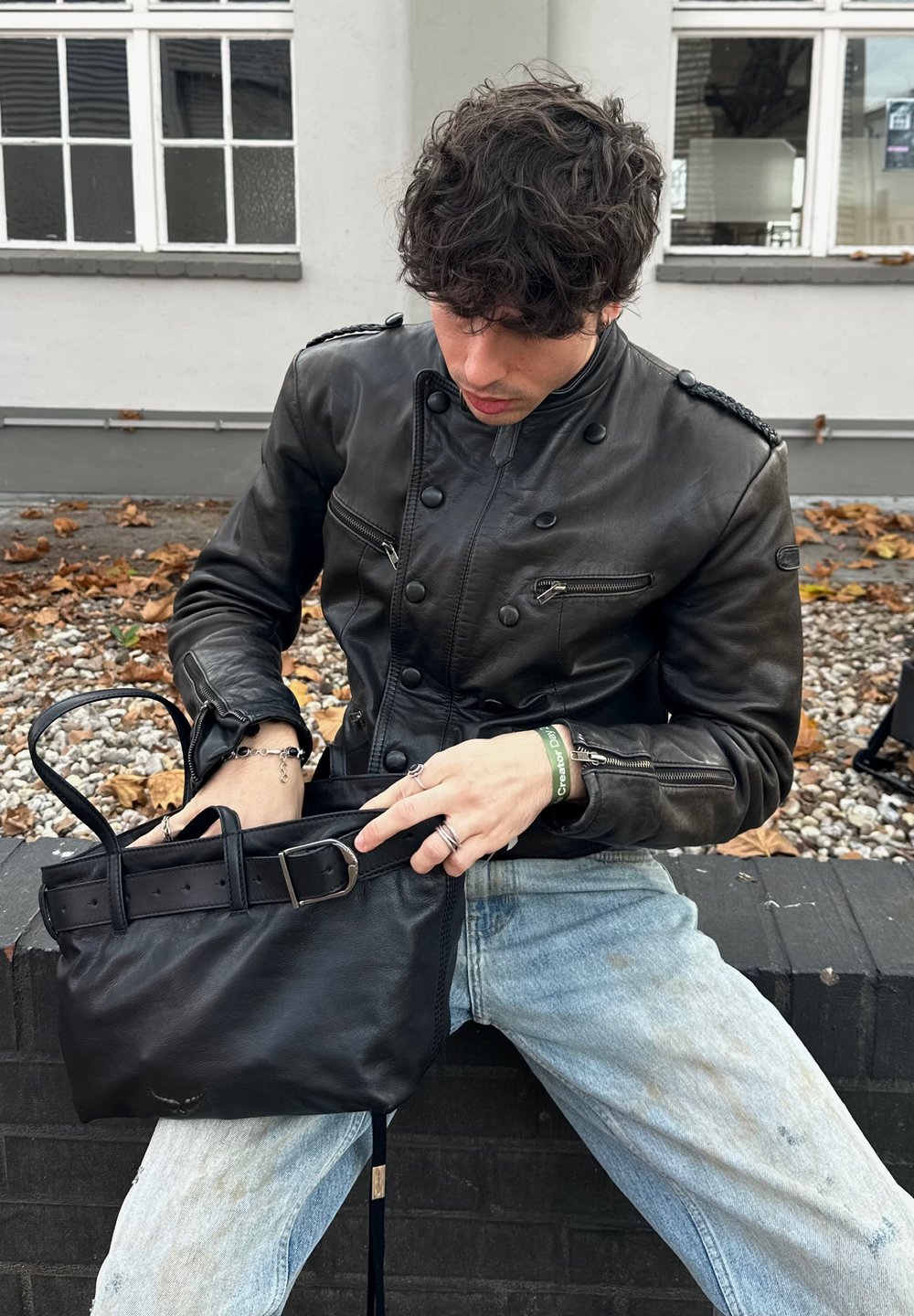 Person med krøllet mørkt hår iført en sort læderjakke og lyseblå jeans sidder på en lav mur og leder i en sort håndtaske.