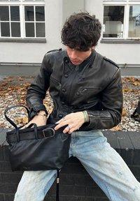 Person med krøllet mørkt hår iført en sort læderjakke og lyseblå jeans sidder på en lav mur og leder i en sort håndtaske.