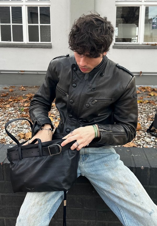 Person med krøllet mørkt hår iført en sort læderjakke og lyseblå jeans sidder på en lav mur og leder i en sort håndtaske.