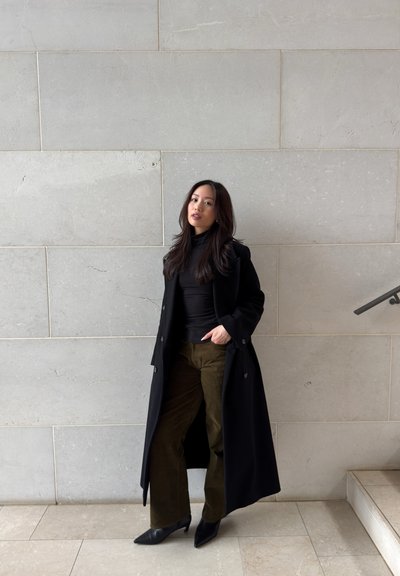 Mujer con cabello largo y oscuro que lleva un abrigo negro, top negro, pantalones color oliva y tacones negros, de pie contra una pared de piedra clara cerca de unas escaleras.