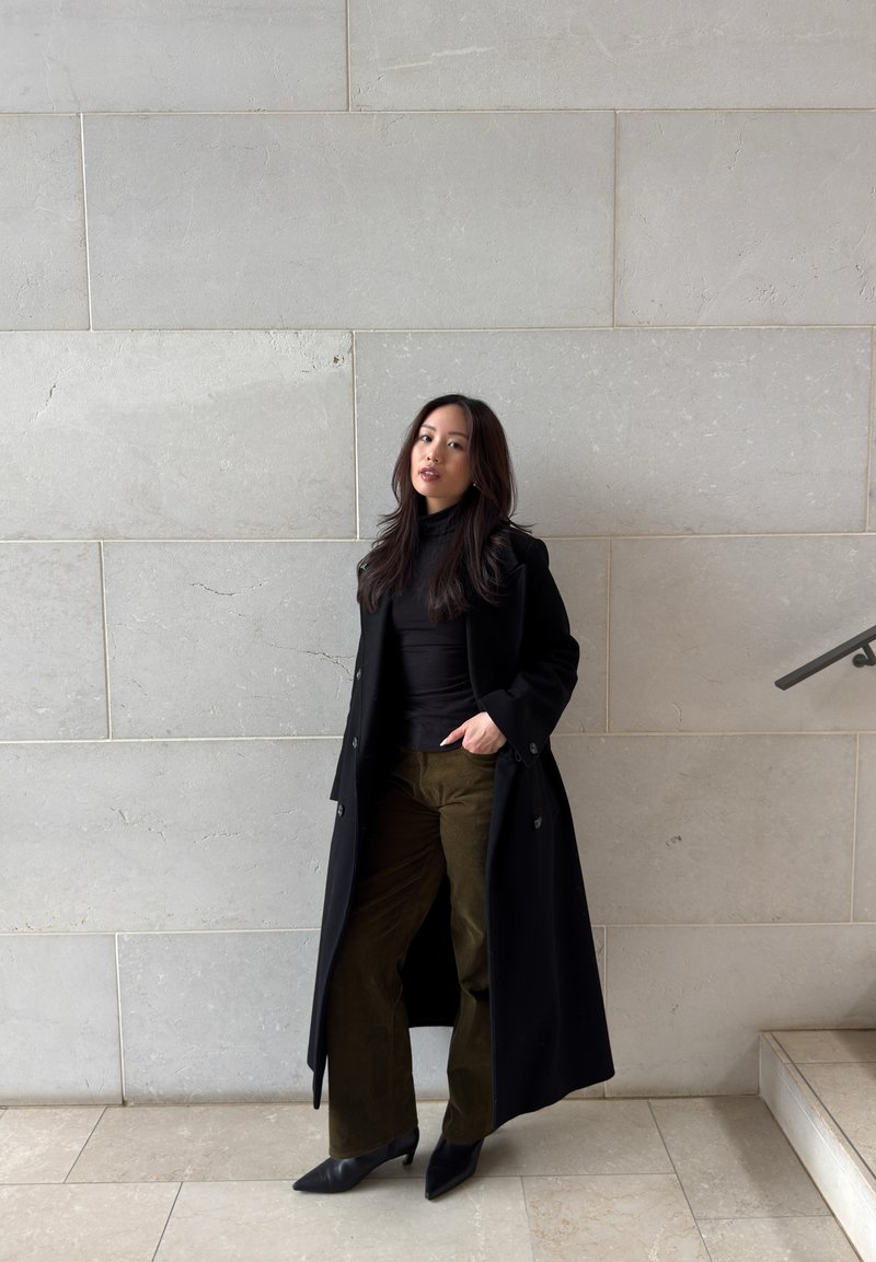 Femme aux longs cheveux foncés portant un manteau noir, un haut noir, un pantalon olive et des talons noirs, debout contre un mur en pierre claire près des escaliers.