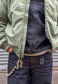 Rohelise bomber-tüüpi jakk ribadega mansettide ja tumeda teksaste overalliga. Võtmehoidja, millel on helmed ja võtmed, ripub taskust.