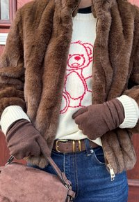 Casaco de pele sintética castanho com um suéter creme que apresenta um estampado de um urso vermelho. Luvas castanhas, um cinto de camurça castanho e uma bolsa a condizer completam o visual.