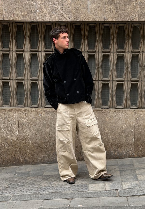 Jeune homme debout sur un trottoir carrelé, portant une veste noire, un pantalon cargo beige large et des chaussures brunes, devant un mur de pierre texturé avec des fenêtres géométriques.