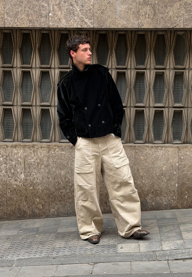Jeune homme debout sur un trottoir carrelé, portant une veste noire, un pantalon cargo beige large et des chaussures marron, devant un mur de pierre texturé avec des fenêtres géométriques.