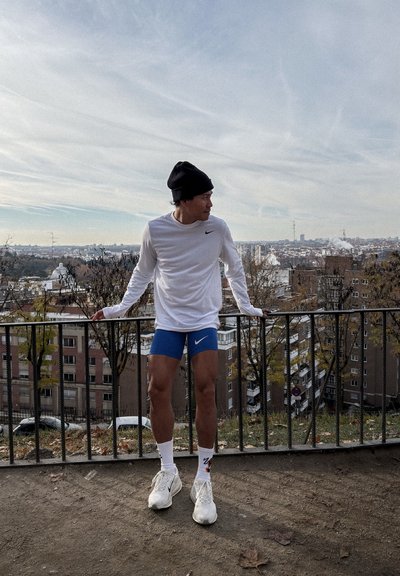 Hombre con camisa blanca de manga larga, pantalones cortos azules, zapatillas blancas y gorro negro apoyado en una barandilla de metal con vista a un paisaje urbano bajo un cielo nublado.
