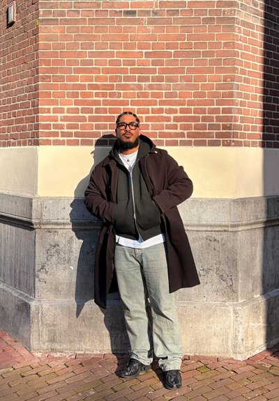 Hombre con gafas y cabello trenzado de pie con las manos en los bolsillos contra una pared de esquina de ladrillo y piedra, vistiendo abrigo, sudadera con capucha, vaqueros y zapatillas.