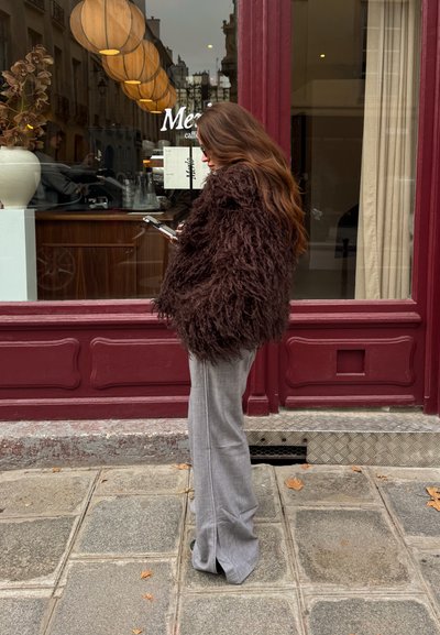 Abrigo de piel sintética marrón y peludo, llevado sobre pantalones acampanados de color gris claro; de pie frente a una cafetería con detalles de madera y luces colgantes redondas.