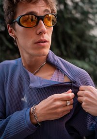 Jong persoon met kort haar en oranje zonnebril die een paarse, gestructureerde jas openritst, met een ketting, armbanden en een ring buiten draagend.