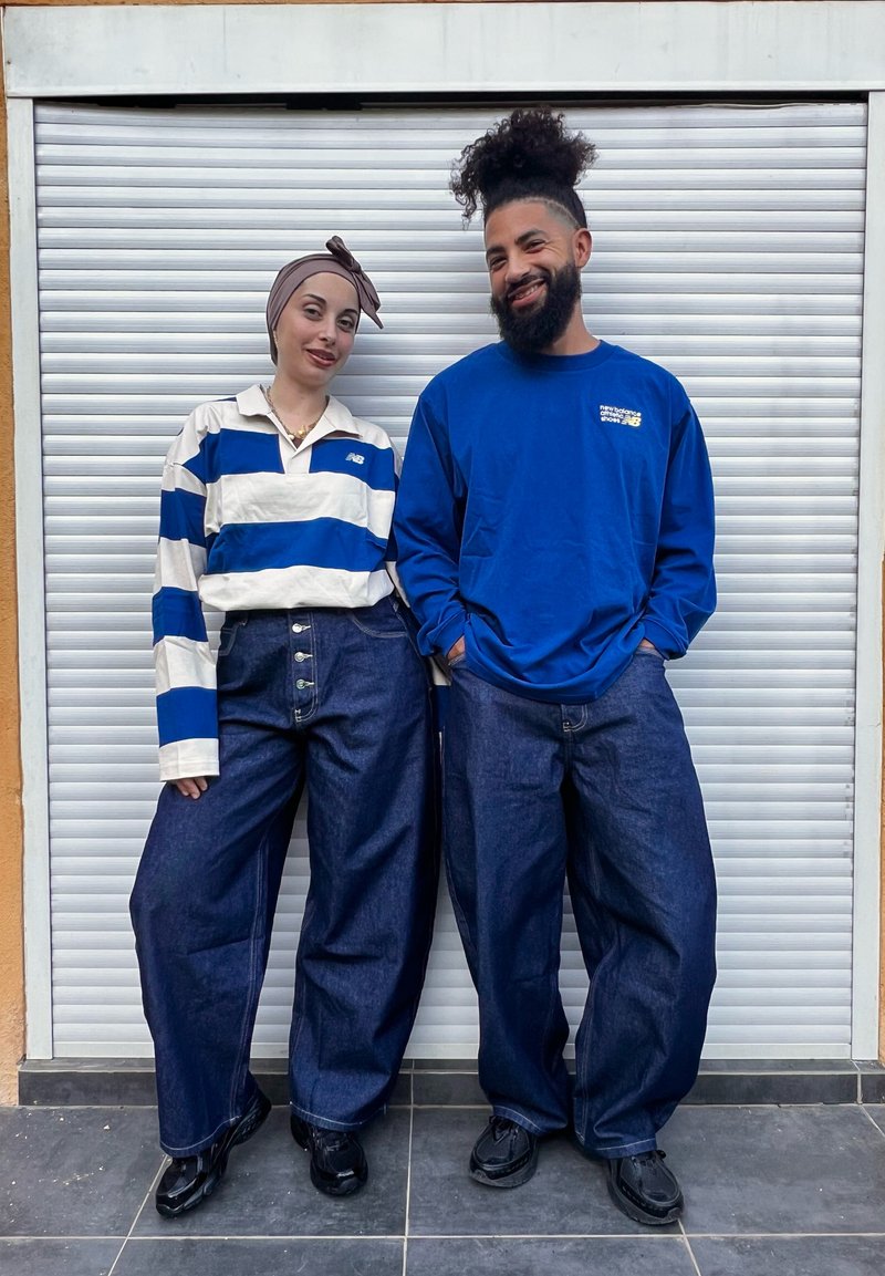 Duas pessoas estão de pé contra um fundo fechado. Uma veste uma polo azul e branca com riscas, e a outra uma camisa azul lisa de manga longa. Ambas usam calças de ganga de perna larga e sapatos pretos elegantes.