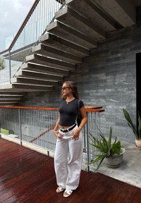 Svart tajt t-shirt, vita vida byxor och vita sandaler. Betongtrappa och stenvägg i en modern miljö med grönska.