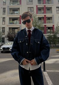 Dżinsowa kurtka z srebrnymi guzikami, dużymi przednimi kieszeniami, w luźnym kroju, połączona z białą koszulą i ciemnym krawatem. Czarne okulary przeciwsłoneczne dopełniają stylizację.