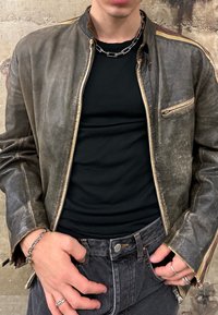 Jachetă de bomber din piele maro cu fermoar atât pe față cât și pe laterale, având un contur bej, textură uzată și detalii minime din metal. Cămașă neagră dedesubt.