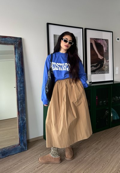 Mujer con cabello largo y oscuro, usando gafas de sol, sudadera azul, falda beige, calcetines con estampado animal y zapatillas, de pie en el interior cerca de una obra de arte enmarcada.