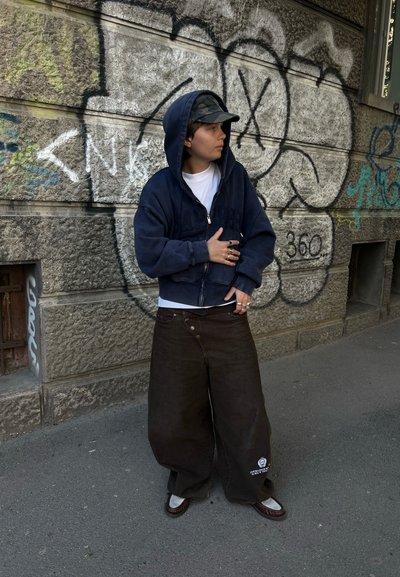 Persona con una sudadera con capucha azul marino, gorra de camuflaje y pantalones anchos oscuros, de pie junto a una pared de piedra cubierta de grafitis en una acera de la ciudad.