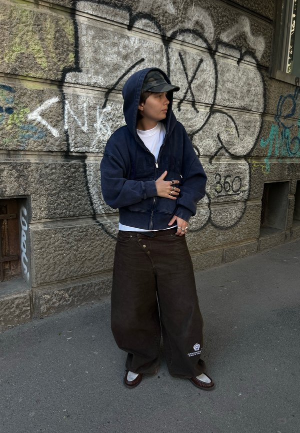 Henkilö, jolla on päällään tummansininen huppari, maastokuvioinen lippis ja leveät tummat housut, seisoo graffitein peitetyn kiviseinän vieressä kaupungin jalkakäytävällä.