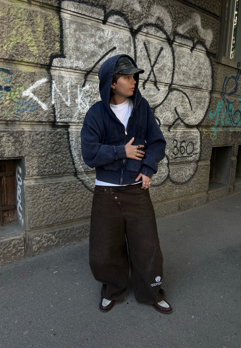 Personne portant un sweat à capuche bleu marine, une casquette camouflage et un pantalon large et sombre se tient contre un mur en pierre couvert de graffiti sur un trottoir en ville.