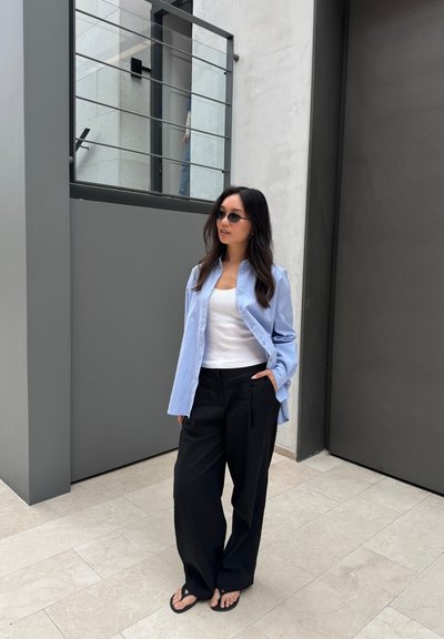 Camisa de botones azul claro, top de tirantes blanco acanalado, pantalones de pierna ancha negros y chanclas negras, situados en un espacio arquitectónico moderno.