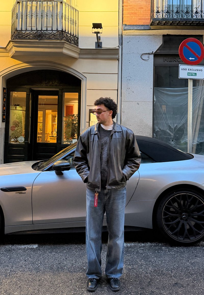 Muž s brýlemi, koženou bundou a džínami stojí s rukama v kapsách vedle stříbrného kabrioletu na městské ulici blízko hotelu.