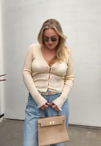 Γυναίκα με ξανθά μαλλιά που φοράει γυαλιά ηλίου, ένα μπεζ ριμπ πλεκτό ζακετάκι, μπλε τζιν και κρατά μια μπεζ υφασμάτινη τσάντα μπροστά σε ένα απλό τοίχο.