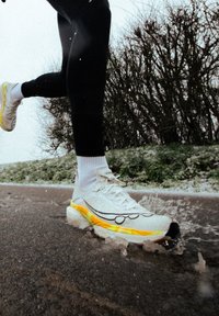 Personne portant des chaussures de course blanches et jaunes éclaboussant de l'eau en courant sur un sentier extérieur humide avec des arbres nus en arrière-plan.