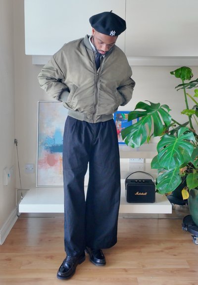 Hombre con boina negra, chaqueta bomber oliva, pantalones anchos negros y zapatos negros, de pie en el interior cerca de una planta y una obra de arte.