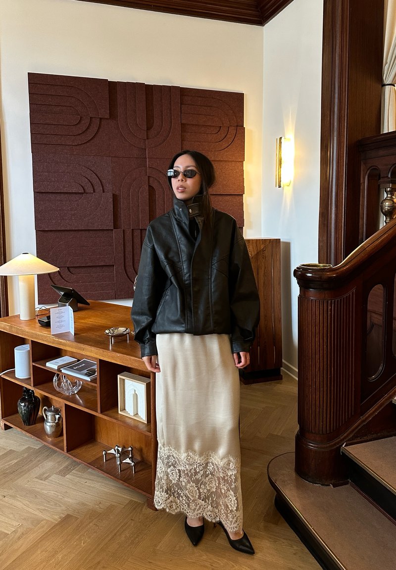 Donna con giacca di pelle nera, gonna beige con bordo in pizzo e occhiali da sole scuri, in piedi accanto a una scrivania in legno e una scala in un interno moderno.