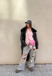 Jeune femme portant un manteau noir, une chemise rose, un pantalon gris usé, une casquette grise et un sac bandoulière rose, debout contre un mur beige.