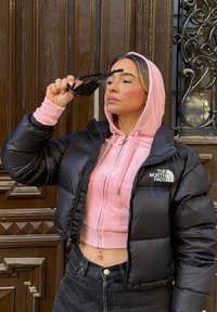 Musta toppatakki, jossa kiiltävä kangas, vaaleanpunainen lyhyt huppari alla, mustat farkut, aurinkolasit kädessä, seisoo puuoven edessä.