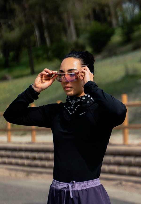 Vrouw die buiten een paar paars getinte zonnebrillen aanpast, draagt een zwart topje met lange mouwen, een patroon nekwarmer en paarse broek met trekkoord.