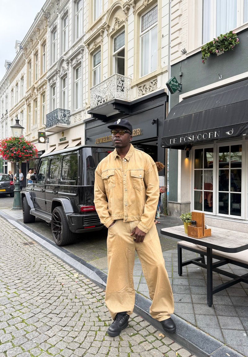 Beige denimjacka med två bröstfickor och matchande vida byxor. Svarta sneakers och keps. Urban gatumiljö med affärer och en svart SUV.