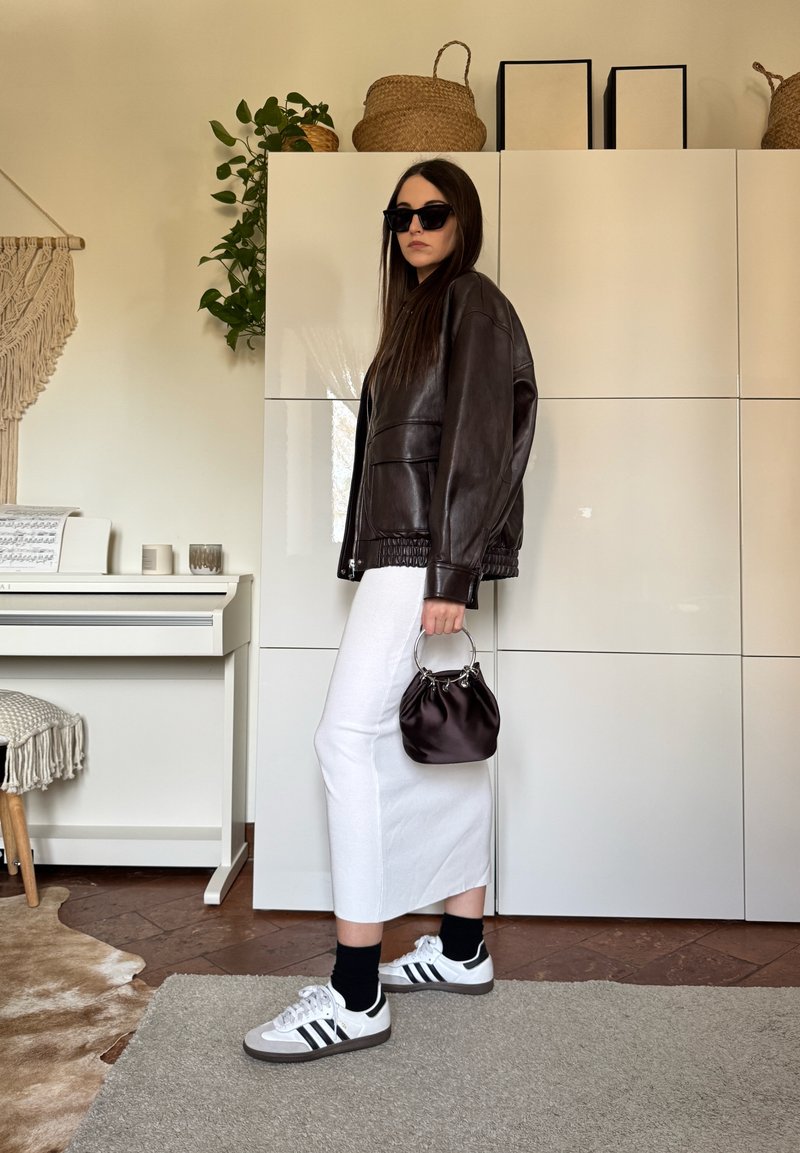 Femme portant une veste en cuir noire, une jupe blanche, des lunettes de soleil noires et des baskets, tenant un petit sac à main noir dans une pièce moderne.