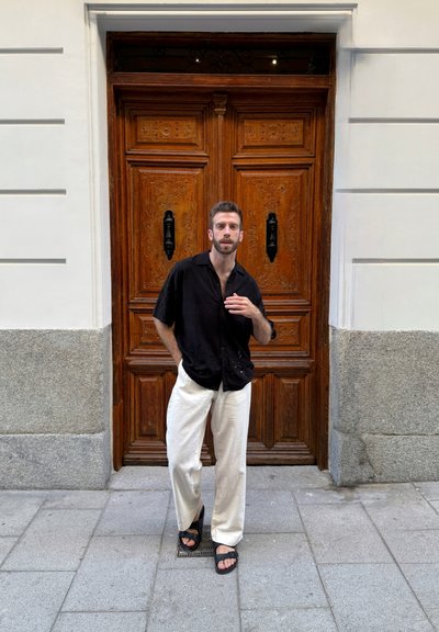 Camisa negra de manga corta, pantalones color crema y sandalias negras llevadas frente a una puerta de madera ornamentada con tallados decorativos y manijas negras.