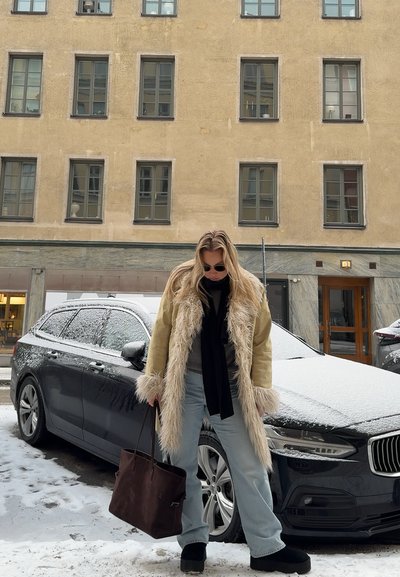 Abrigo beige forrado de pelo largo, bufanda negra, camiseta gris, jeans celestes y zapatos de plataforma negros. Bolsa tote marrón oscuro. Calle y coche cubiertos de nieve.
