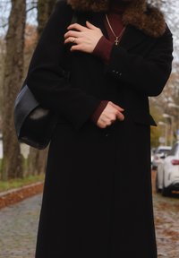 Black coat with a fur collar and button details, worn over a burgundy top. A black bag with a textured finish is slung over the shoulder.