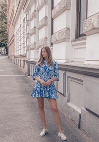 Kleid mit blau-weißen Mustern, 3/4-Ärmeln und einem ausgestellten Rock. Getragen mit weißen Schuhen, vor einem urbanen Hintergrund mit strukturierten Wänden.