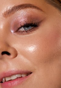 Nærbilde av et ansikt med glatt, strålende hud. Har skinnende rosa øyenskygge, mørke vipper og myk, naturlig leppefarge.