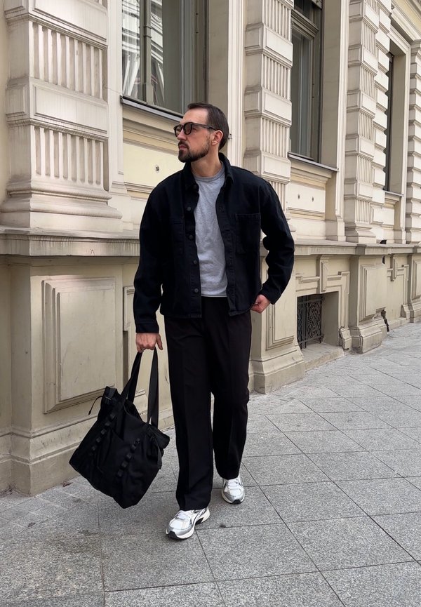 Homme portant une veste et un pantalon noirs, une chemise grise, des baskets blanches, portant un sac fourre-tout noir, marchant sur le trottoir de la ville à côté d'un bâtiment orné.