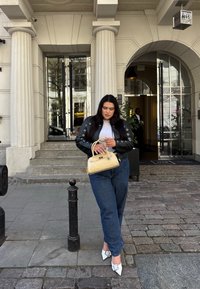 Čierna kožená bunda s očkovými akcentmi, šedý top, modré nohavice s vysokým pásom, biele lodičky s ostro špicatou špičkou a svetložltá kabelka, vonku pri budove.